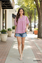 Brunette model walking in Umgee raspberry striped babydoll top with cream crossbody bag and denim cutoff shorts, outdoor boutique lifestyle shot