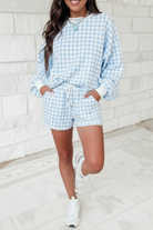Sky blue gingham drawstring shorts with white elastic waistband, hands in pockets, white brick wall lifestyle