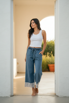 Full-length lifestyle shot of brunette woman in wide-leg washed denim pants with floral lace ankle trim standing in a bright airy doorway, white ribbed tank and gold jewelry — BiBi lace trim pants