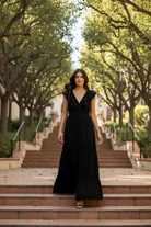 Brunette model walking outdoors in a long black ruffle tiered maxi dress with deep V-neckline and ruffle cap sleeves, full body lifestyle shot