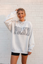 Oversized gray PRAY embroidered sweatshirt with polka dot lettering, laughing lifestyle pose, white stone wall background