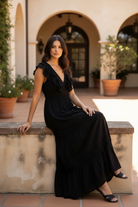 Brunette model seated on outdoor steps in long black tiered ruffle maxi dress, relaxed pose showing full skirt length in golden afternoon light