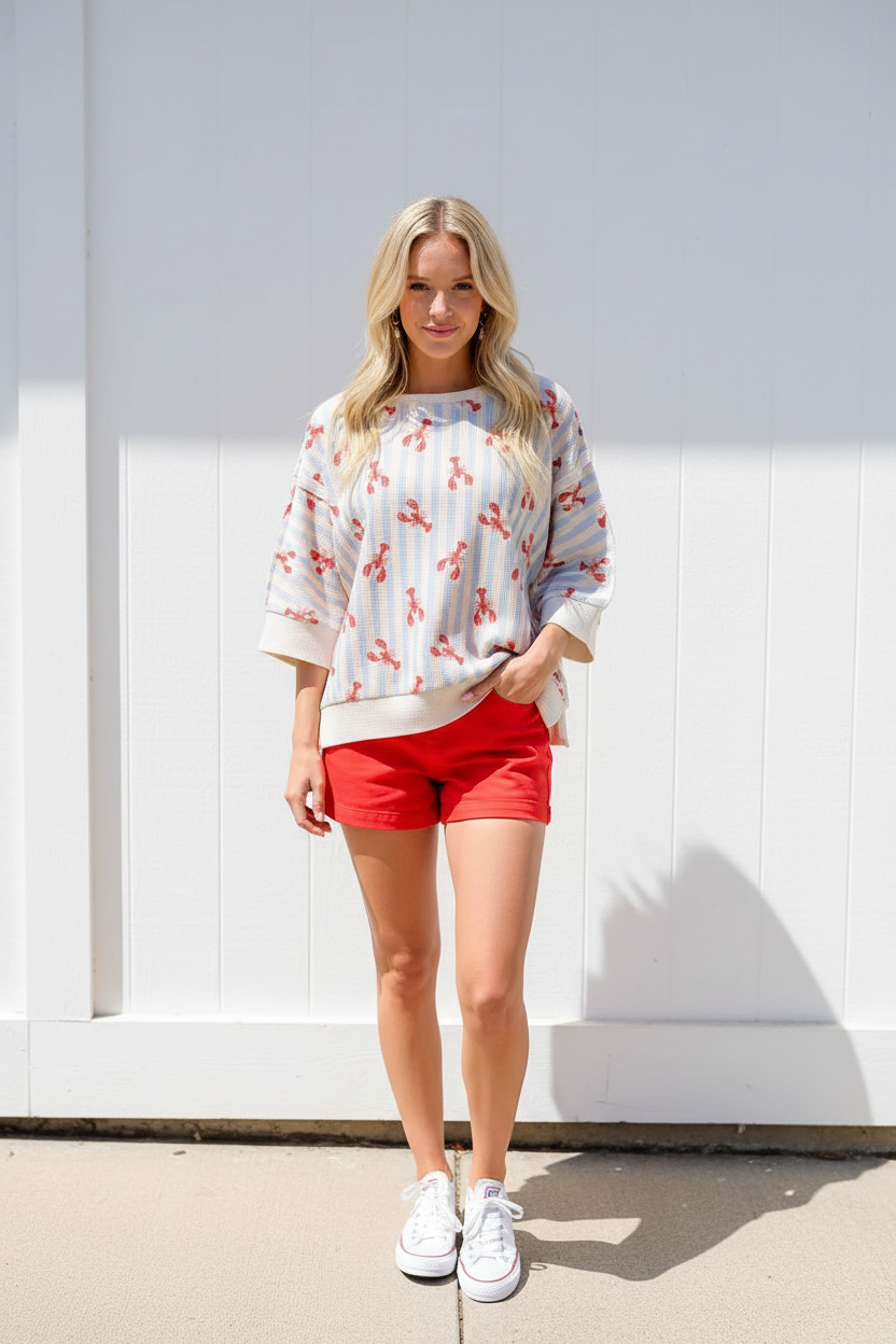 Blonde woman in a light blue and red lobster stripe top with waffle knit sleeves and red shorts, standing against a white wall in bright summer sunlight — festive 4th of July outfit