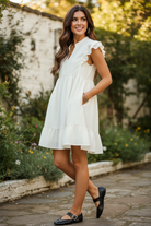 Cream textured woven ruffle sleeve babydoll mini dress three-quarter lifestyle view showing double-layer ruffle cap sleeves, gathered empire waist, full skirt silhouette, and single ruffle tier hi-low hem in outdoor garden setting
