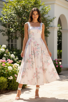 Brunette model wearing blush floral square neck open back midi dress with wide tie straps and full A-line pleated skirt, front-facing lifestyle pose in sunlit garden patio