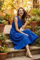 Royal blue smocked waist tiered midi dress seated lifestyle — brunette model on garden steps tiers fanned out courtyard terracotta pots