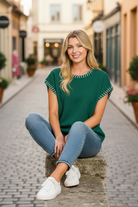 Hunter green oversized short sleeve knit sweater outdoor seated lifestyle photo, relaxed casual pose, white whipstitch detail, soft cotton blend, styled with denim jeans