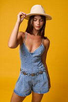 Woman wearing a light wash denim romper with deep V neckline, tie-back detail, and pleated shorts styled with a cream hat and coin belt on yellow background
