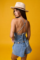 Woman wearing a light wash denim romper with deep V neckline, tie-back detail, and pleated shorts styled with a cream hat and coin belt on yellow background