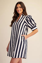 A woman modeling a short puff-sleeve shirt dress with vertical navy and white stripes, set against a plain beige studio background.