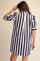 A woman shown from the back wearing a short puff-sleeve shirt dress with bold vertical navy and white stripes, set against a plain beige studio background.
