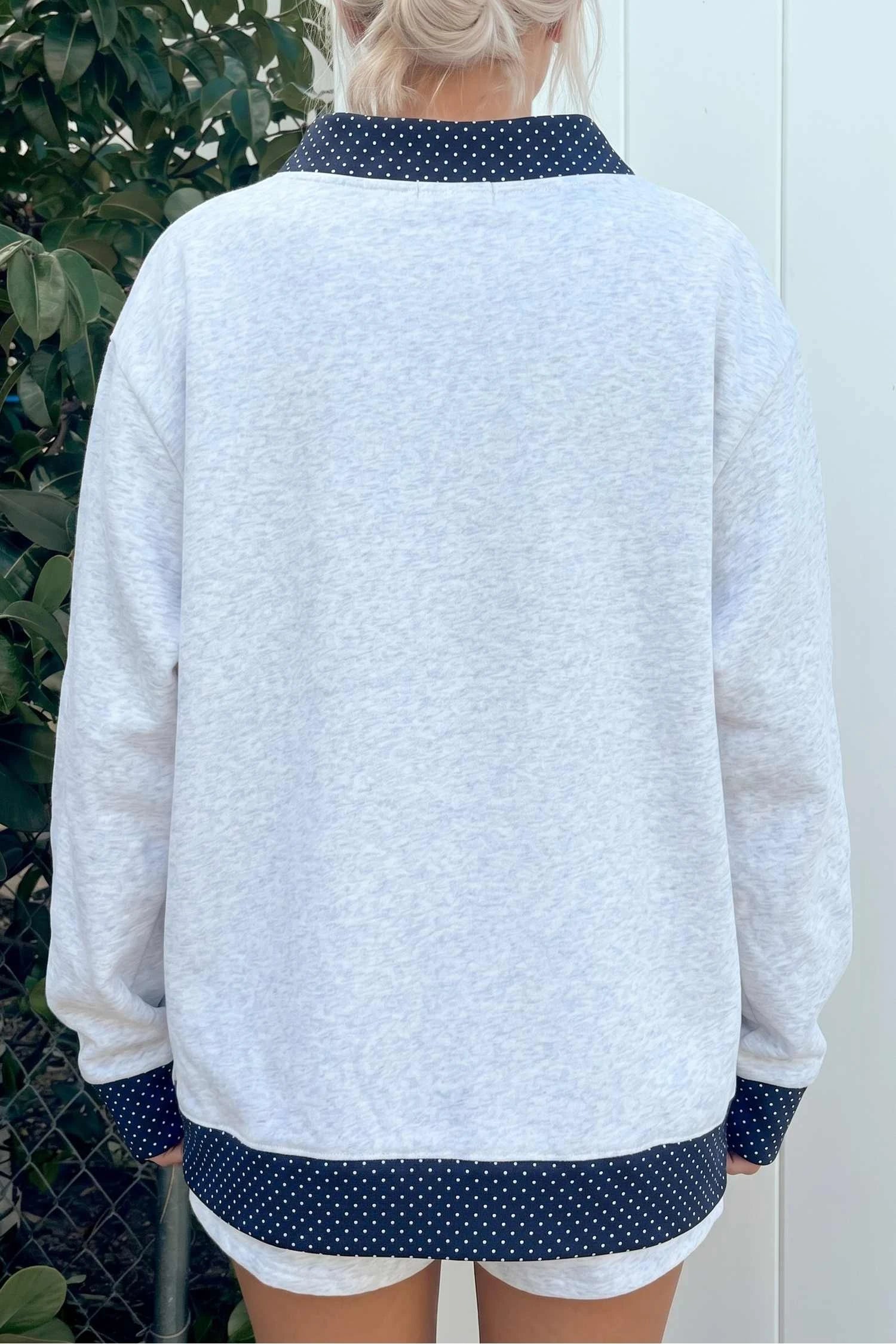 A person shown from the back wearing a light heather gray sweatshirt with dark navy polka-dot trim on the collar, cuffs, and hem, paired with white shorts.