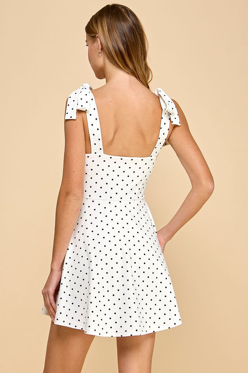 A smiling blonde woman wears a white polka-dotted fit-and-flare dress with bow-tie shoulder straps. The dress features a sweetheart neckline, built-in lined shorts, and a lightweight, breathable fabric, blending feminine style with athleisure-inspired comfort.