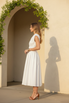 Side profile of brunette model in white zip front smocked waist midi dress highlighting cinched waist silhouette and flowing A-line skirt, Mediterranean archway, boutique lifestyle photo